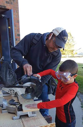 A man and young boy wearing safety glasses work together to cut a board with a chop saw. 